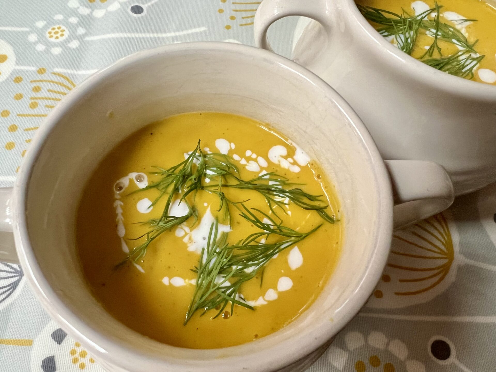 Two bowls of creamy butternut squash, carrot soup garnished with swirls of cream and fresh dill, placed on a patterned tablecloth.