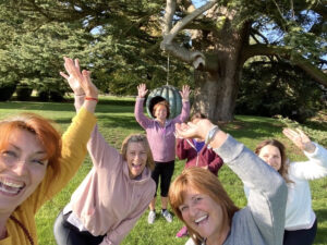 Five women pose outdoors, smiling and waving at the camera in front of a large tree and a tire swing on a sunny day.