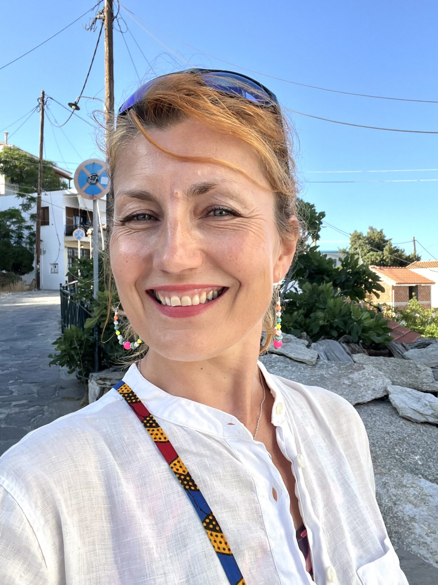 A woman with light brown hair and colorful earrings smiles outdoors on a sunny day, wearing a white blouse. There are buildings, plants, and a blue sky in the background.
