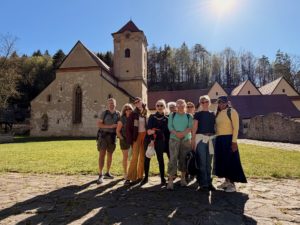 Eight people stand together posing for a photo in front of an old stone church with a tall tower, surrounded by trees and buildings, on a sunny day.