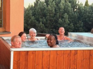 Five adults are sitting and smiling in an outdoor hot tub with bubbling water, against a backdrop of green trees and an orange building wall.