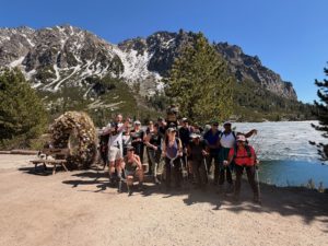 A group of hikers pose on a dirt path by a lake with mountains and pine trees in the background; a flower-covered arch and a picnic table are visible on the left.