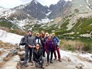 A group of eight people poses on a rocky path in a mountainous landscape with patches of snow, greenery, and a small lake.