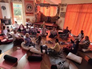 A group of people sit in a circle on cushions and yoga mats in a sunlit room, appearing to participate in a group meditation or wellbeing session.