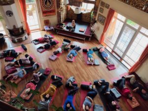 A group of people practise yoga on mats in a spacious room with wooden floors, large windows, and colourful decor. An instructor sits on a platform at the front.